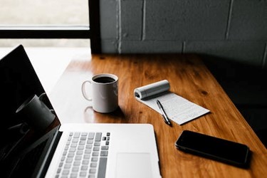 laptop open on desk with coffee, pen, paper and mobile to hand