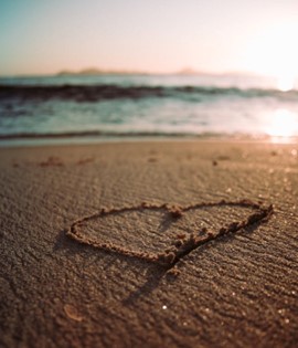Heart drawn into sand on beach, sun glistening on the water