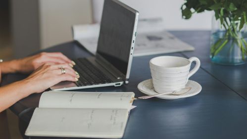 hands typing on laptop next to cup and open diary