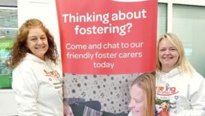 two foster carers by pull up stand with text " Thinking about fostering? Come and chat to our friendly foster carers today